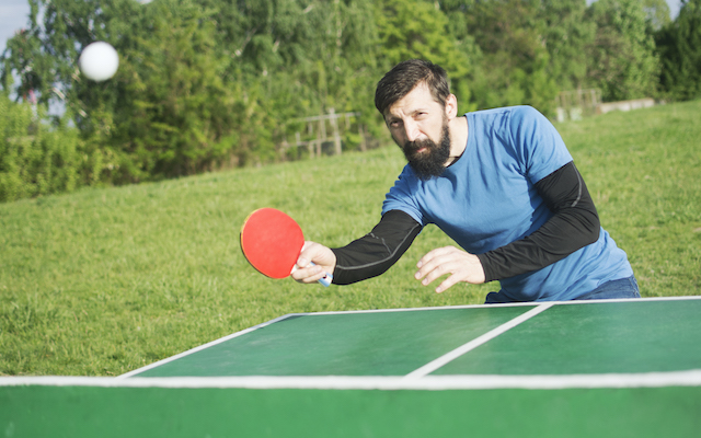 The bearded man is playing table tennis or pingpong outdoors as recreation.
