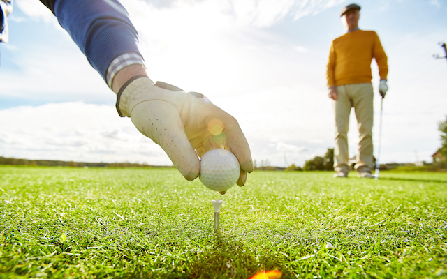 One of golf players putting ball on tee while the other one standing on background