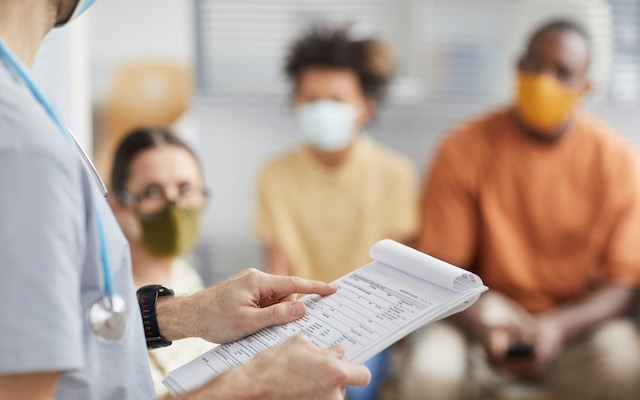 A clinician stands with a clipboard in front of people wearing Covid-19 masks.
