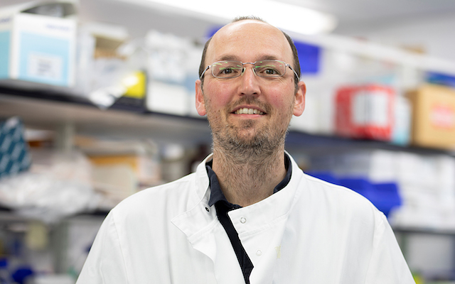 Research scientist Andy Howden smiles for a photo in a lab coat.
