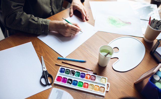 A person paints at a table filled with art supplies.