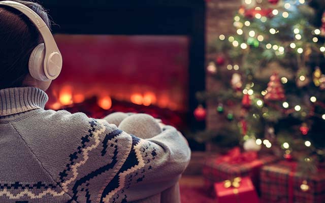 A man sits in front of a fireplace and Christmas tree with headphones on.