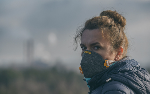 Woman wearing a mask standing in front a city backdrop showing smoke and pollution