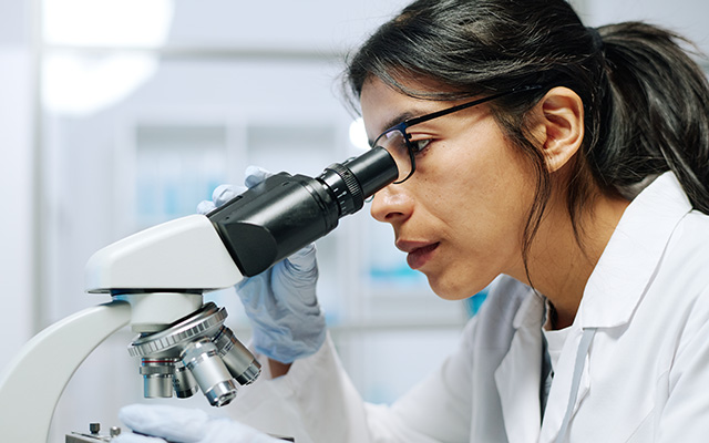 Side view of young serious female biochemist in whitecoat, gloves and eyeglasses looking in microscope during scientific research