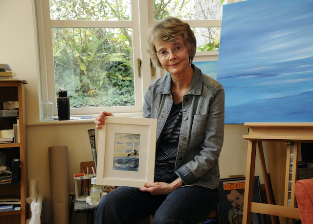 Sitting in an art studio, Kathleen Reardon displays a painting of a lighthouse