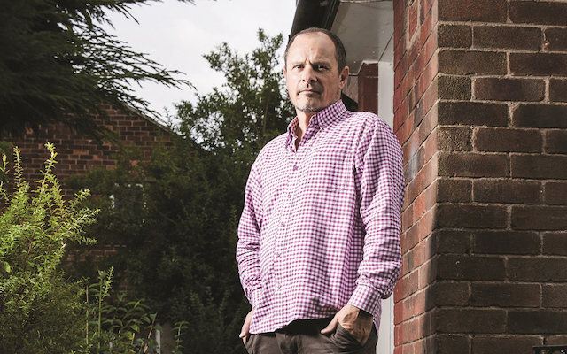 Matt Eagles stands outside beside a brick wall and a bush, posing for a photo.