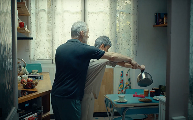 Screenshot from 'living with Parkinson's disease' featuring man standing behind a woman helping her pour coffee