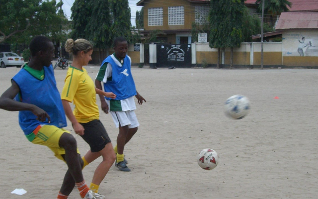 Tania Park wearing a yellow t-shirt playing football with two men wearing blue t-shirts